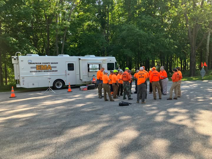 Searchers in orange Search & Rescue shirts and helmets receive a briefing beside the Champaign County Sheriff EMA mobile command vehicle.