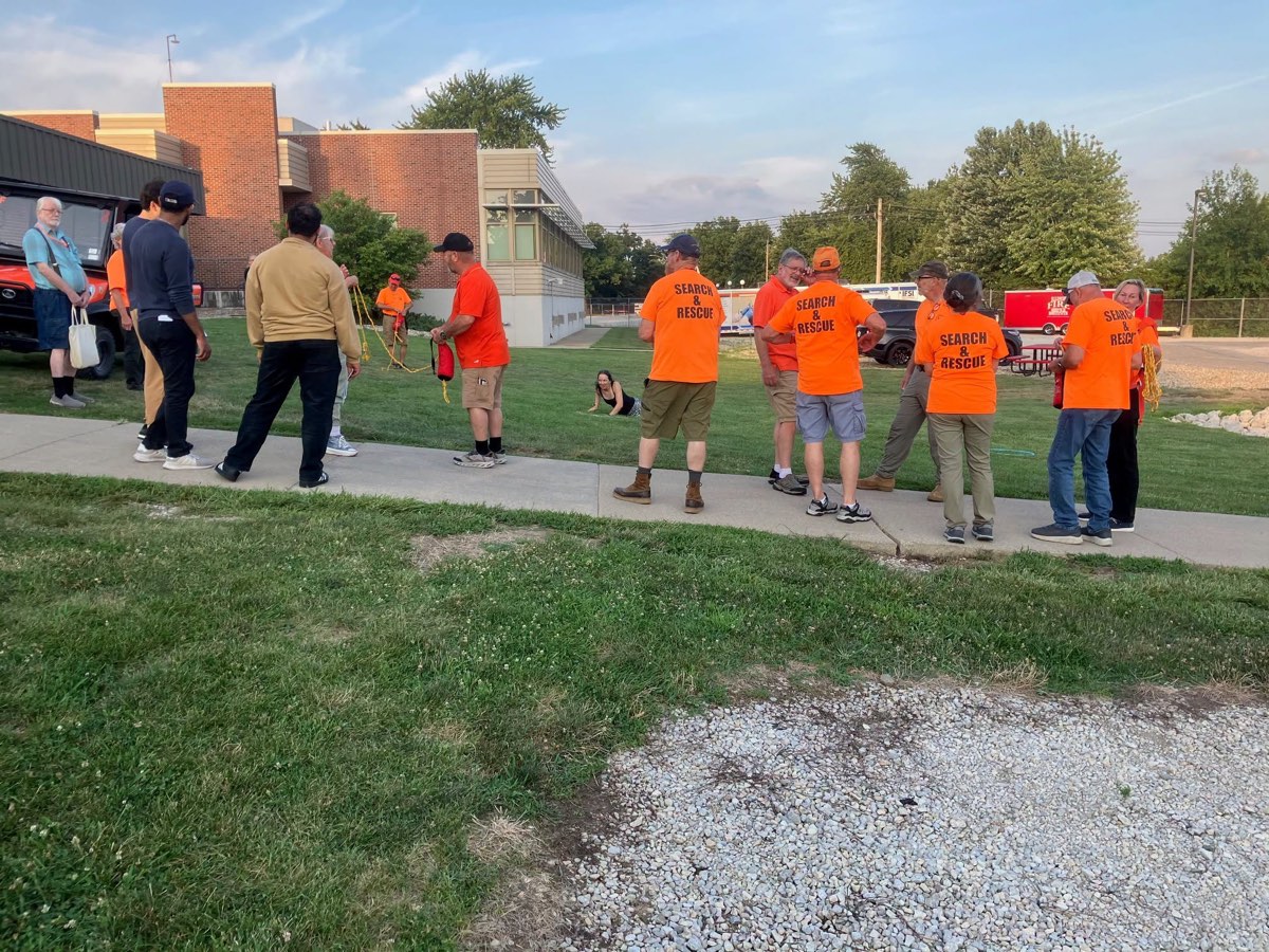 Team members in orange Search & Rescue shirts stand on the lawn outside IFSI coiling throw bags, with a training target seated on the grass ahead of them.