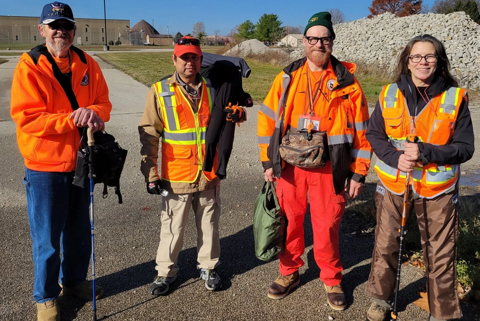Four volunteers wearing orange jackets, reflective vests, and field gear stand together outdoors.