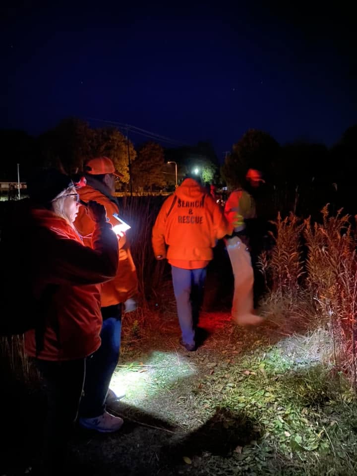 Searchers in orange jackets work along a brushy field edge after dark, lighting the ground with headlamps and flashlights.
